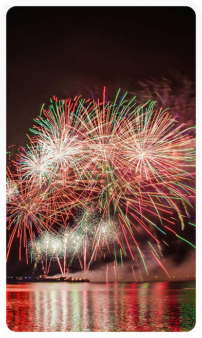 Colorful fireworks bursting over calm water at night with reflections on the surface.
